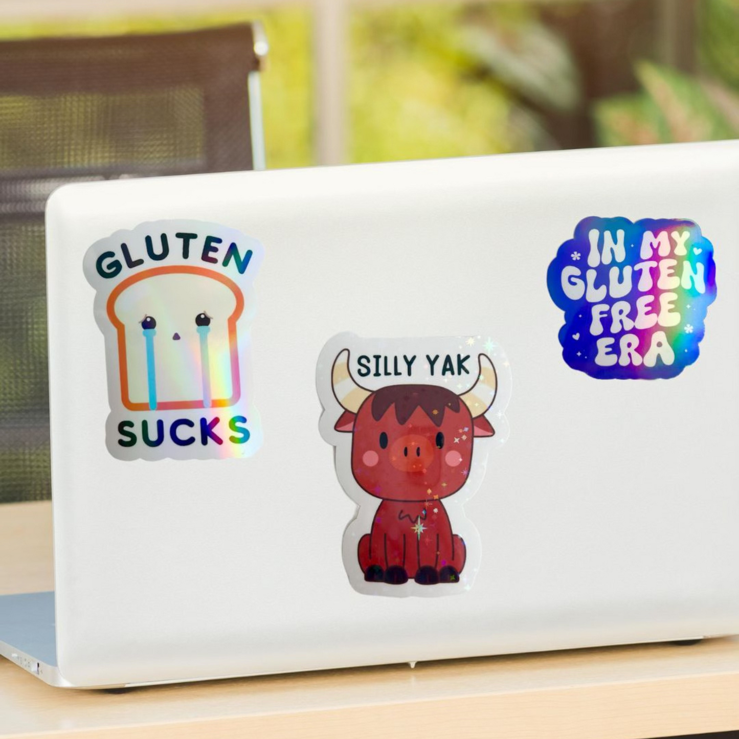White laptop decorated with three holographic gluten free stickers, including “Gluten Sucks,” “Silly Yak,” and “In My Gluten Free Era” designs.
