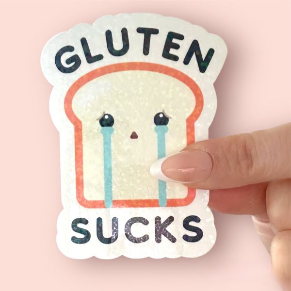Hand holding a “Gluten Sucks” sticker with a crying toast character and holographic finish.
